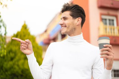 Kafkasyalı genç bir adam açık havada kahvesini yudumlarken yan tarafı göstererek bir ürün sunuyor.