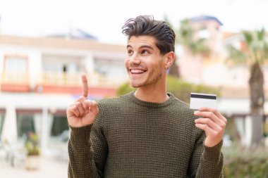 Açık havada kredi kartı tutan genç beyaz bir adam çözümün farkına varmak için parmağını kaldırıyor.