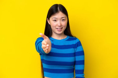 Sarı arka planda izole edilmiş genç bir Çinli kadın iyi bir anlaşma için el sıkışıyor.