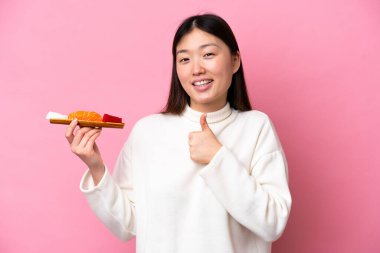 Elinde saşimi tutan genç Çinli kadın pembe arka planda izole edilmiş bir şekilde baş parmak işareti yapıyor.