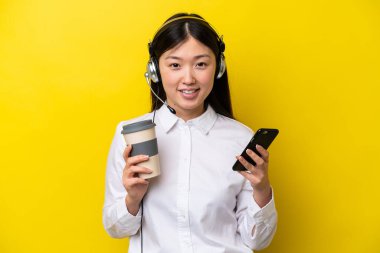 Tele pazarlamacı Çinli bir kadın sarı arka planda izole edilmiş bir kulaklıkla çalışıyor. Elinde kahve ve cep telefonu var.