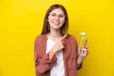 Sarı arka planda izole edilmiş su şişesi olan genç bir İngiliz kadın bir ürünü sunmak için kenarı işaret ediyor.