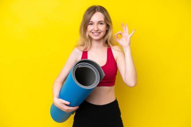 Genç beyaz kadın yoga derslerine giderken sarı arka planda izole edilmiş bir minder tutuyor. Parmaklarıyla tamam işareti gösteriyor.