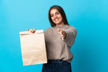 Mavi arka planda izole edilmiş bir paket yiyecek alan bir kadın iyi bir anlaşma için el sıkışıyor.