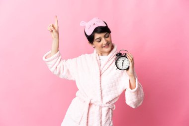 Kısa saçlı bir kadın pembe arka planda pijamalı ve mutlu bir ifadeyle saati tutuyor.