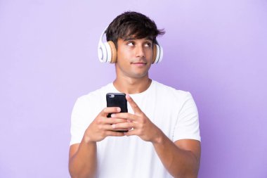 Young caucasian man isolated on purple background listening music with a mobile and thinking