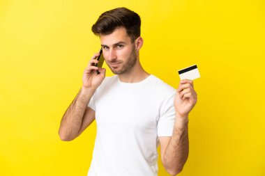 Sarı arka planda izole edilmiş genç beyaz adam cep telefonuyla sohbet ediyor ve elinde bir kredi kartı tutuyor.