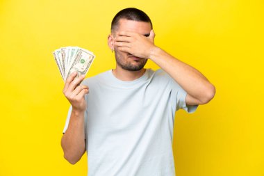 Kafkas 'lı genç bir adam, sarı arka planda izole edilmiş ve gözleri elle kapatılmış bir sürü para alıyor. Bir şey görmek istemiyorum.