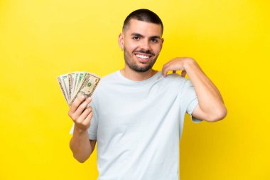 Kafkasyalı genç bir adam, sarı arka planda izole edilmiş bir sürü para alıyor ve gülüyor.