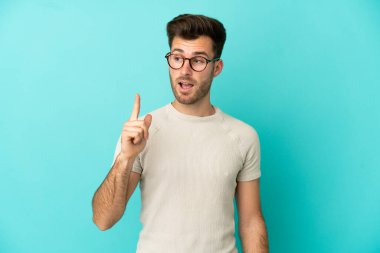 Genç, beyaz, yakışıklı bir adam mavi arka planda izole edilmiş. Çözümü bulmak için parmağını kaldırıyor.
