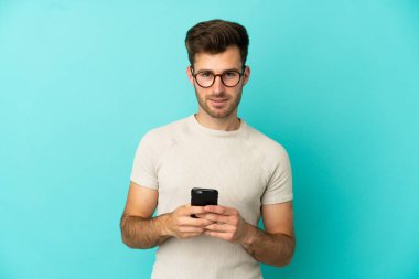 Mavi arka planda izole genç beyaz adam cep telefonuyla bir mesaj gönderiyor.