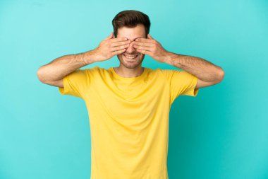 Beyaz tenli yakışıklı adam mavi arka planda izole edilmiş gözleri elleriyle kaplamış ve gülümsüyor.