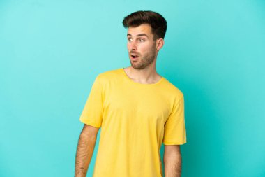 Genç, beyaz, yakışıklı bir adam mavi arka planda izole edilmiş. Yan tarafa bakarken sürpriz jest yapıyor.