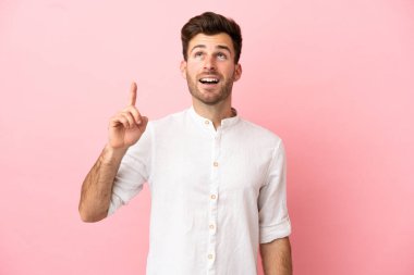 Pembe arka planda izole edilmiş beyaz, yakışıklı genç bir adam yukarı bakıyor ve şaşırıyor.