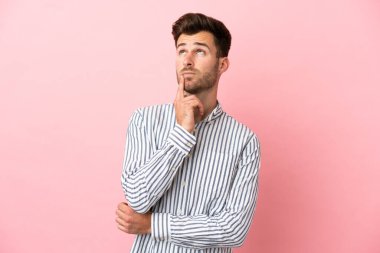 Pembe arka planda izole edilmiş beyaz, yakışıklı genç bir adam yukarı bakarken şüphe duyuyor.
