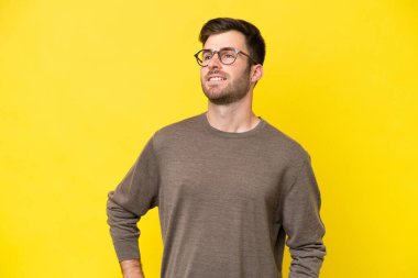 Sarı arka planda izole edilmiş genç beyaz adam kolları kalçasında ve gülümsüyor.