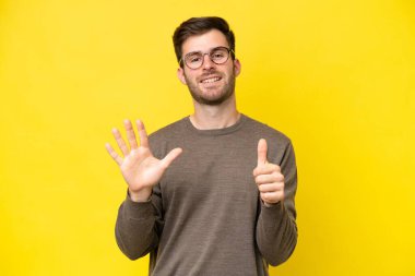 Sarı arka planda izole edilmiş genç beyaz adam parmaklarıyla altısını sayıyor.