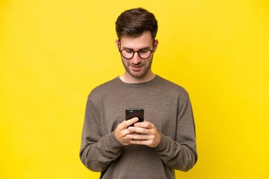 Sarı arka planda izole edilmiş genç beyaz adam cep telefonuyla bir mesaj gönderiyor.