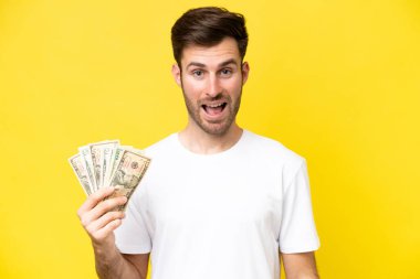Kafkasyalı genç bir adam, sarı arka planda, şaşırtıcı bir yüz ifadesiyle izole edilmiş bir sürü para alıyor.
