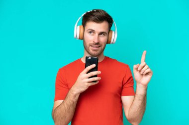 Kafkasyalı genç adam mavi arka planda izole edilmiş. Cep telefonuyla müzik dinliyor ve şarkı söylüyor.