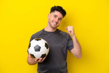 Yakışıklı genç futbolcu sarı arka planda izole edilmiş bir şekilde zaferi kutluyor.