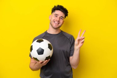 Yakışıklı genç futbolcu sarı arka planda izole edilmiş gülümsüyor ve zafer işareti gösteriyor.