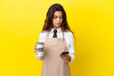 Kafkas lokantası garsonu sarı arka planda izole edilmiş. Elinde kahve ve cep telefonu var.