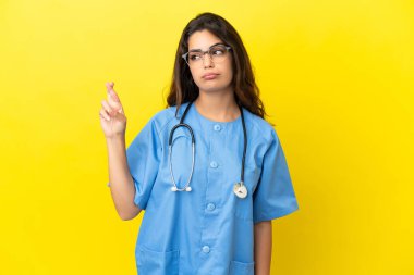 Sarı arka planda izole edilmiş genç cerrah kadın parmaklarını çapraz yapıp en iyisini diliyor.