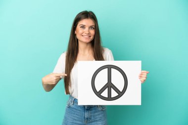 Kafkasyalı genç bir kadın mavi arka planda izole edilmiş. Elinde barış sembolü olan bir levha var.