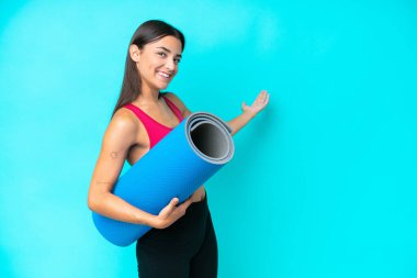Young sport caucasian woman going to yoga classes while holding a mat isolated on blue background extending hands to the side for inviting to come