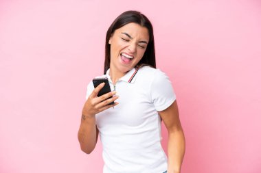 Pembe arka planda izole edilmiş genç beyaz kadın cep telefonu kullanıyor ve zafer jesti yapıyor.