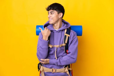 İzole edilmiş sarı arka planı olan büyük bir sırt çantasıyla genç dağcı bir ürünü sunmak için kenarı işaret ediyor.