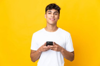 Sarı arka plan izole edilmiş genç adam kameraya bakıyor ve cep telefonunu kullanırken gülümsüyor.
