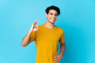 Genç Venezuelalı adam mavi arka planda izole edilmiş. Parmaklarıyla işaret veriyor.