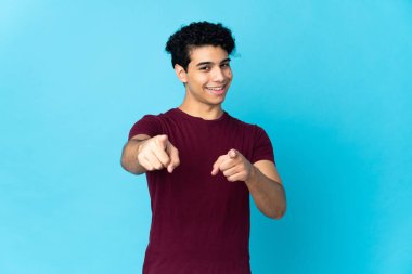 Genç Venezuelalı bir adam mavi arka planda izole edilmiş mutlu bir ifadeyle cepheyi işaret ediyor