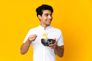 Genç Venezuelalı bir adam sarı arka planda izole edilmiş. Elinde çubuklu erişte kasesi var ve yukarı bakıyor.