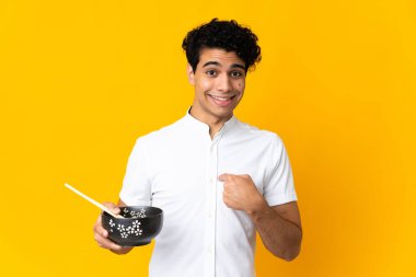 Genç Venezuelalı bir adam, elinde yemek çubuklarıyla bir kase erişte tutarken, sürpriz bir yüz ifadesiyle sarı arka planda izole edilmiş.