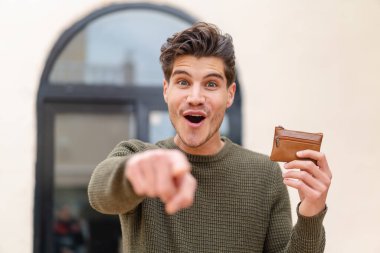 Açık havada cüzdan tutan genç beyaz bir adam şaşırmış ve ön tarafı işaret ediyor.