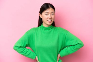 Pembe arka planda izole edilmiş genç Çinli kadın kolları kalçasında ve gülümsüyor.