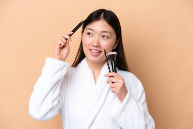 Bej arka planda izole edilmiş genç Çinli kadın elinde makyaj fırçası tutuyor.