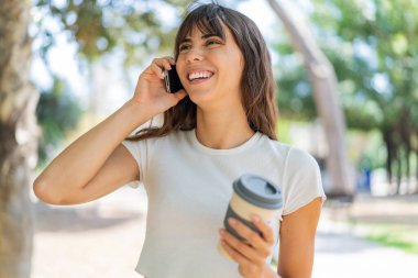 Dışarıda cep telefonu kullanan ve elinde mutlu bir ifadeyle kahve tutan genç bir kadın.
