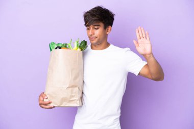Elinde mor arka planda izole edilmiş bir alışveriş çantası tutan genç adam dur hareketi yapıyor ve hayal kırıklığına uğruyor.
