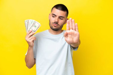 Kafkasyalı genç adam sarı arka planda izole edilmiş bir sürü para alıyor. Durma hareketi yapıyor ve hayal kırıklığına uğruyor.