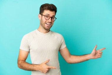 Mavi arka planda izole edilmiş beyaz, yakışıklı genç adam şaşırmış ve işaret ediyor.