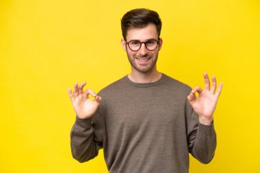 Sarı arka planda izole edilmiş genç beyaz adam iki eliyle tamam işareti gösteriyor.