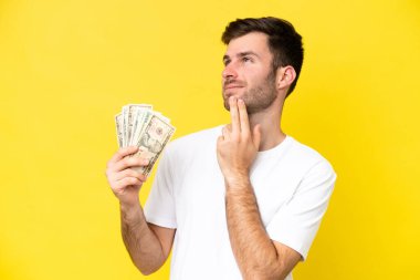 Kafkasyalı genç adam, sarı arka planda izole edilmiş bir şekilde çok para alıyor gülümserken yukarı bakıyor.