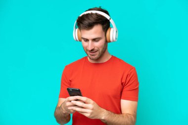 Kafkasyalı genç adam mavi arka planda izole edilmiş müzik dinliyor ve cep telefonu arıyor.