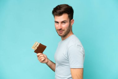 Mavi arka planda izole edilmiş cüzdan taşıyan genç beyaz bir adam çok gülümsüyor.