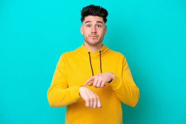 Kafkasyalı genç adam mavi arka planda izole edilmiş. Geç kalma jesti yapıyor.