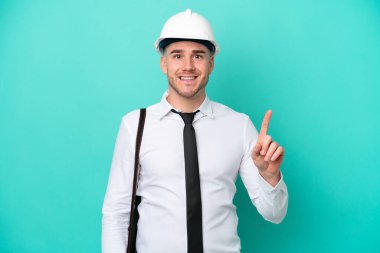 Miğferli ve elinde mavi arka planda izole edilmiş planları olan genç bir beyaz adam en iyinin işareti olarak parmağını kaldırıyor.